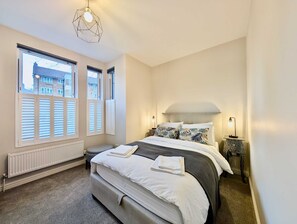 A bedroom with a large bed, a radiator, a window with shutters, and a hanging light fixture. - Stylish 2 Bed Clapham Flat With Private Patio (London)