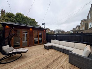 A wooden deck with a sofa and a wicker chair, a wooden structure with glass doors, and a black fence. - Stylish 2 Bed Clapham Flat With Private Patio (London)
