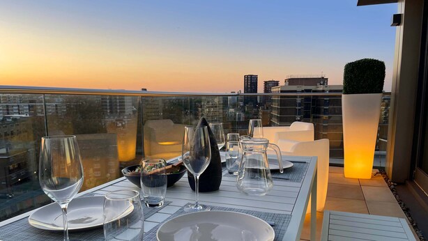 Une table dressée sur un toit, avec des assiettes, des verres et des couverts, d’où l’on peut admirer un panorama urbain au coucher du soleil.