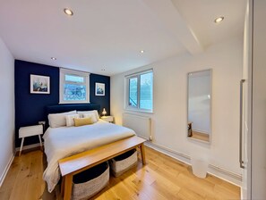 A modern bedroom with a large bed, two bedside tables, a nightstand, a window with blinds, and a radiator. - Stunning London Apartment With Private Patio (London)