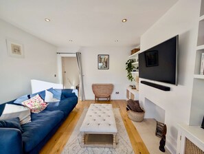 Ein modernes Wohnzimmer mit einem blauen Sofa, einem weißen Hocker, einem Holzstuhl und einem an der Wand befestigten Fernseher.