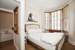 A bedroom with a bed, a bedside table, a mirror, a window with blinds, and a wall-mounted artwork. - Charming Victorian Home in Westminster (London)