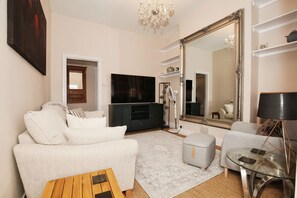 A living room with a sofa, ottoman, coffee table, TV, and a large mirror. - Charming Victorian Home in Westminster (London)