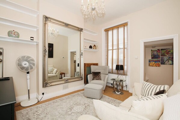 A living room with a large mirror, a fan, a sofa, and a window with blinds. - Charming Victorian Home in Westminster (London)