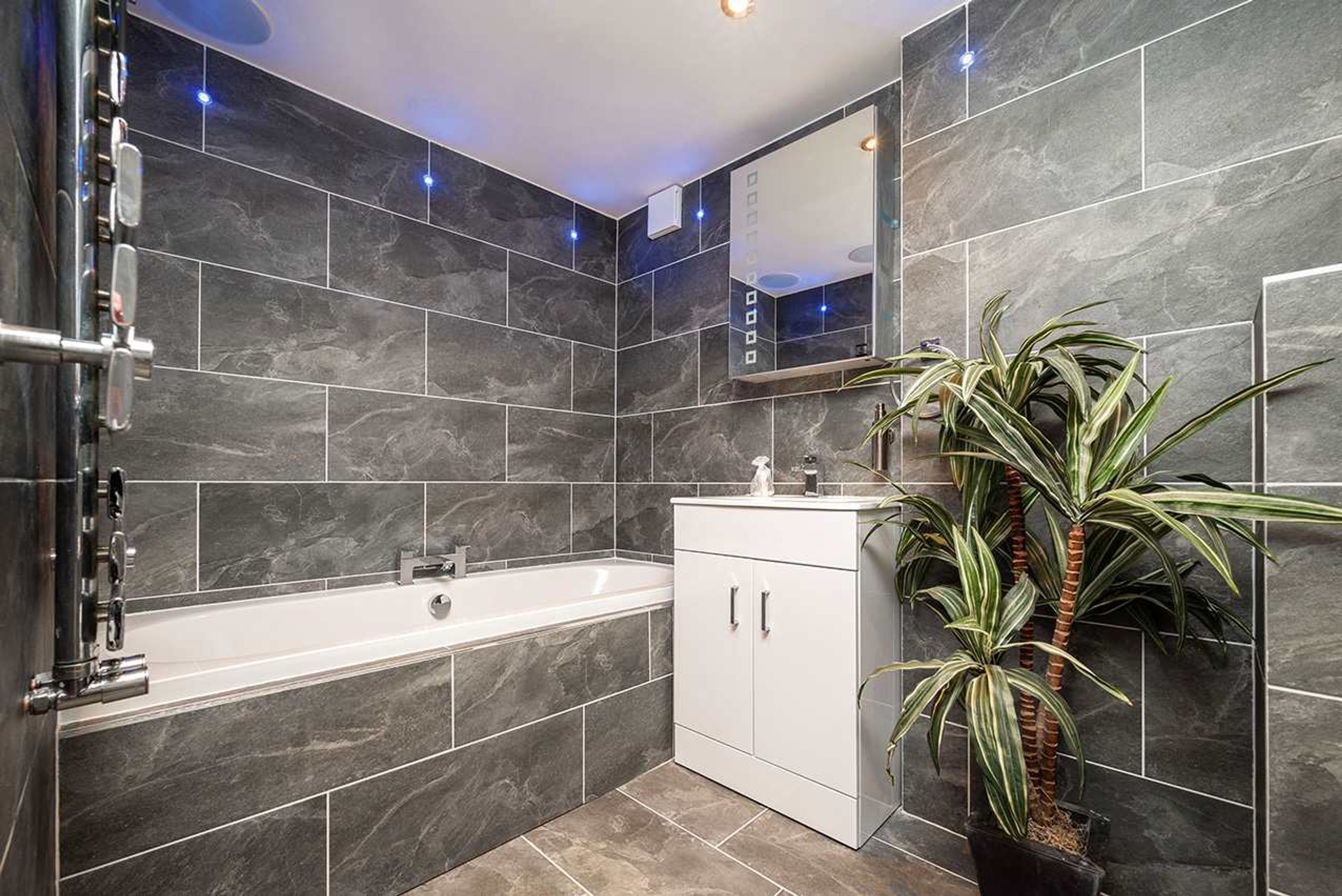 A modern bathroom with dark marble tiles, a white bathtub, a sink with a mirror, and a potted plant.