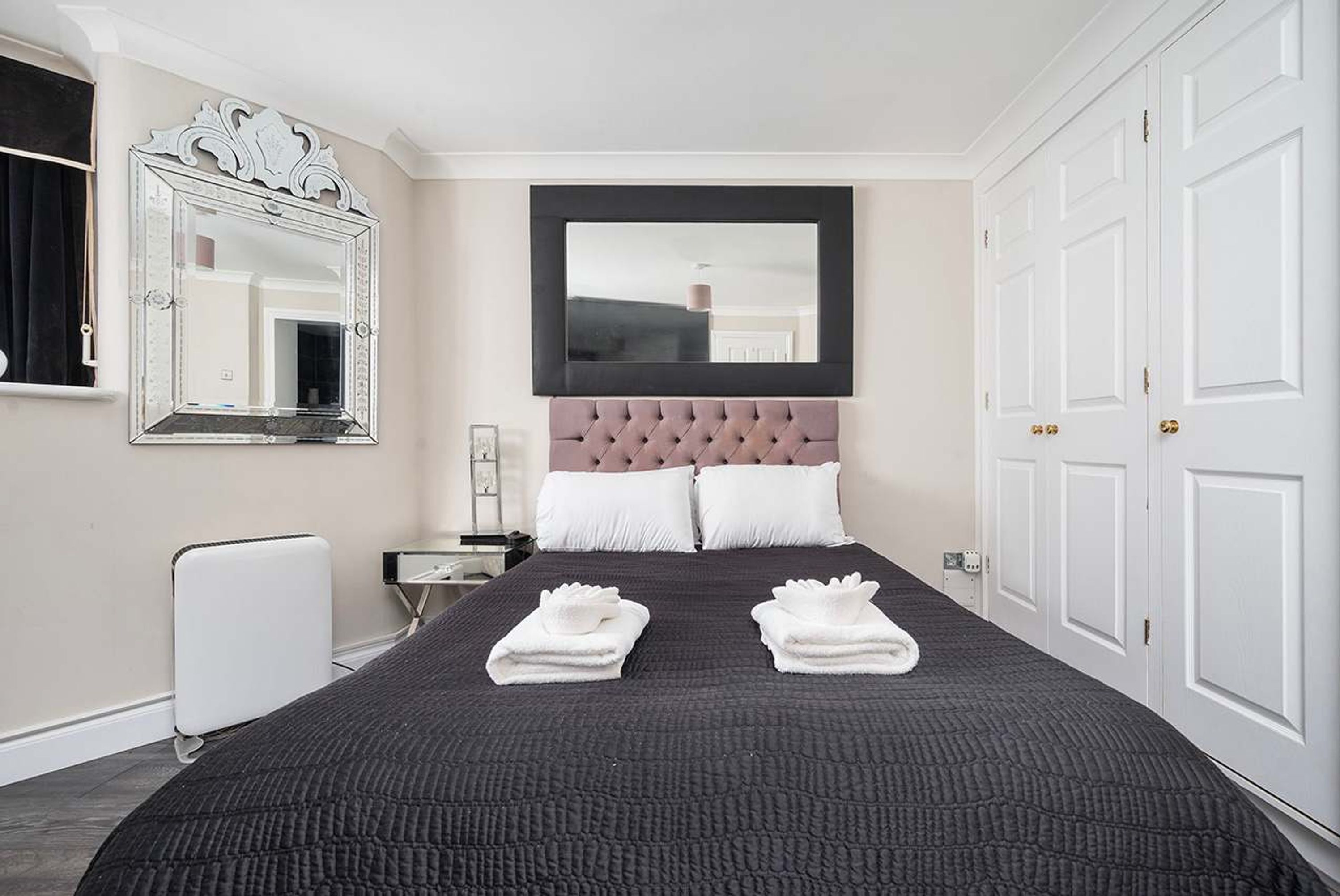 Habitación de hotel con una cama grande, cabecera capitonada, dos almohadas blancas, un espejo amplio y un radiador blanco.