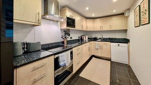 A modern kitchen with light wood cabinets, a black countertop, stainless steel appliances, and recessed ceiling lights. - Victoria Park Retreat Stylish 2 Bed Flat (London)