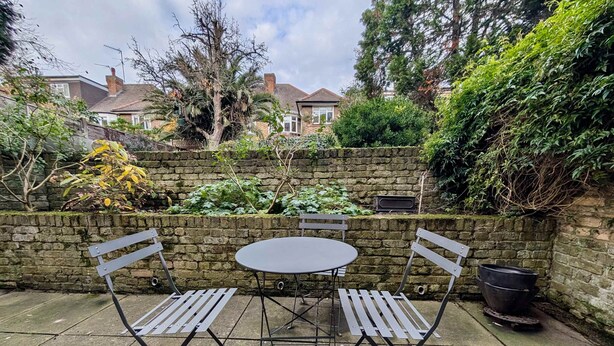 A patio with a round table and two chairs, surrounded by a stone wall and greenery. - Victoria Park Retreat Stylish 2 Bed Flat (London)