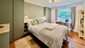 A bedroom with a bed, bedside tables, a desk, a chair, a window with curtains, and a bookshelf. - Victoria Park Retreat Stylish 2 Bed Flat (London)