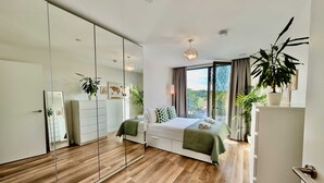 A bedroom with a large mirror, wooden flooring, a bed with green bedding, and a view of greenery outside. - Amazing Flat With Free Parking and Balcony (Woking)
