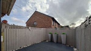 Una terrassa de fusta amb una casa de maó al fons, envoltada per una tanca de fusta i tres jardineres.