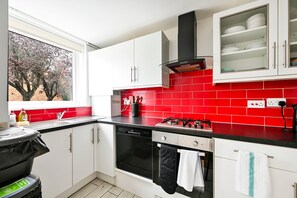 A modern kitchen with white cabinets, a red tiled backsplash, a gas stove, a dishwasher, and a window with a view of trees. - Spacious 5-bedroom Family Home in Greenwhich (London)