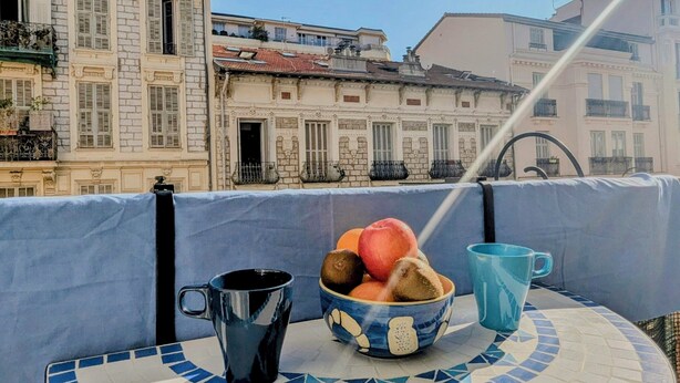 Een tafel met een blauw-wit betegeld patroon, een schaal met fruit en twee kopjes op een balkon met uitzicht op historische gebouwen.