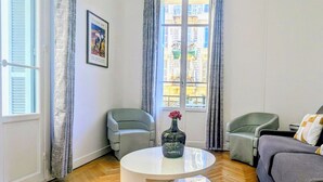 Een moderne woonkamer met een ronde salontafel, twee grijze fauteuils, een bank en een raam met gordijnen.