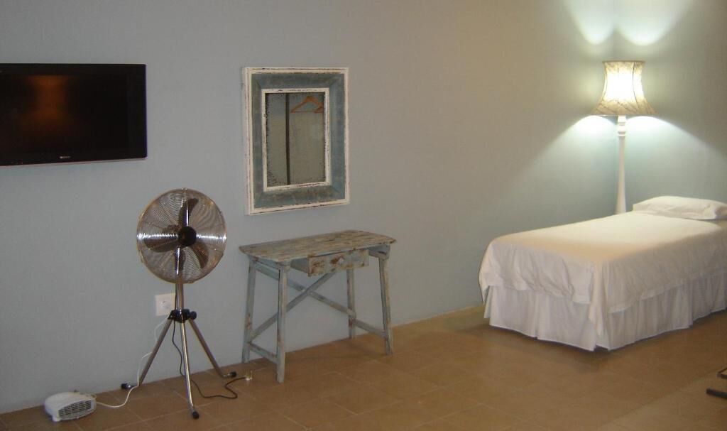 A bedroom with a bed, a small table, a fan, a lamp, and a TV mounted on the wall.