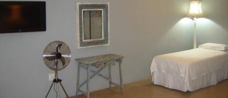 A bedroom with a bed, a small table, a fan, a lamp, and a TV mounted on the wall.