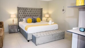 A bedroom with a tufted headboard, a large bed with a white and yellow bedspread, two bedside lamps, a grey bench, and a small potted plant on a shelf. - Rhino Creek Estate (Bloemfontein)