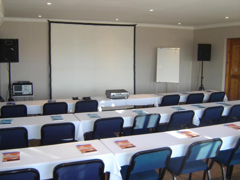 A conference room with rows of tables and chairs, a projector screen, a whiteboard, and audio equipment.
