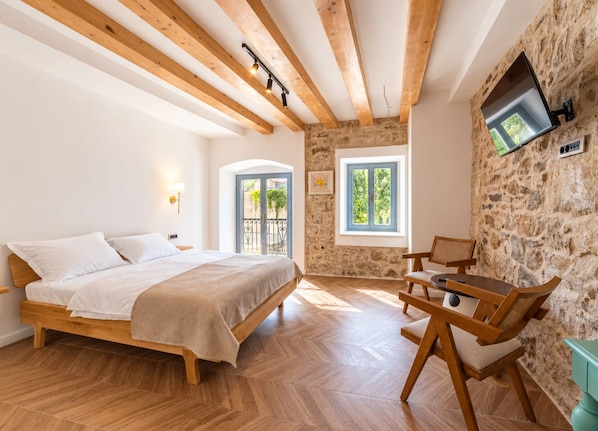 Un dormitorio con cama de madera, pared de piedra, televisor montado en la pared y ventana con vista a vegetación.