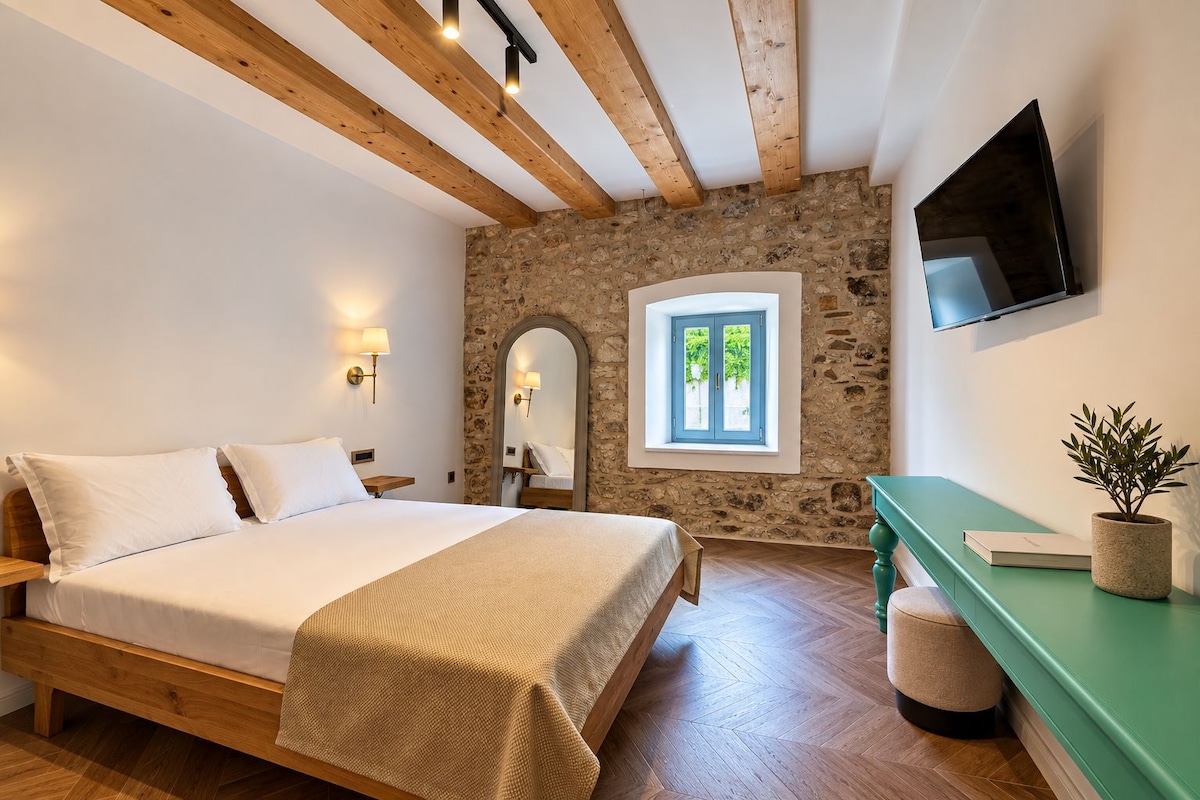 Un dormitorio con cama de madera, pared de piedra, ventana con contraventanas azules, televisor montado en la pared y una consola verde con una planta.