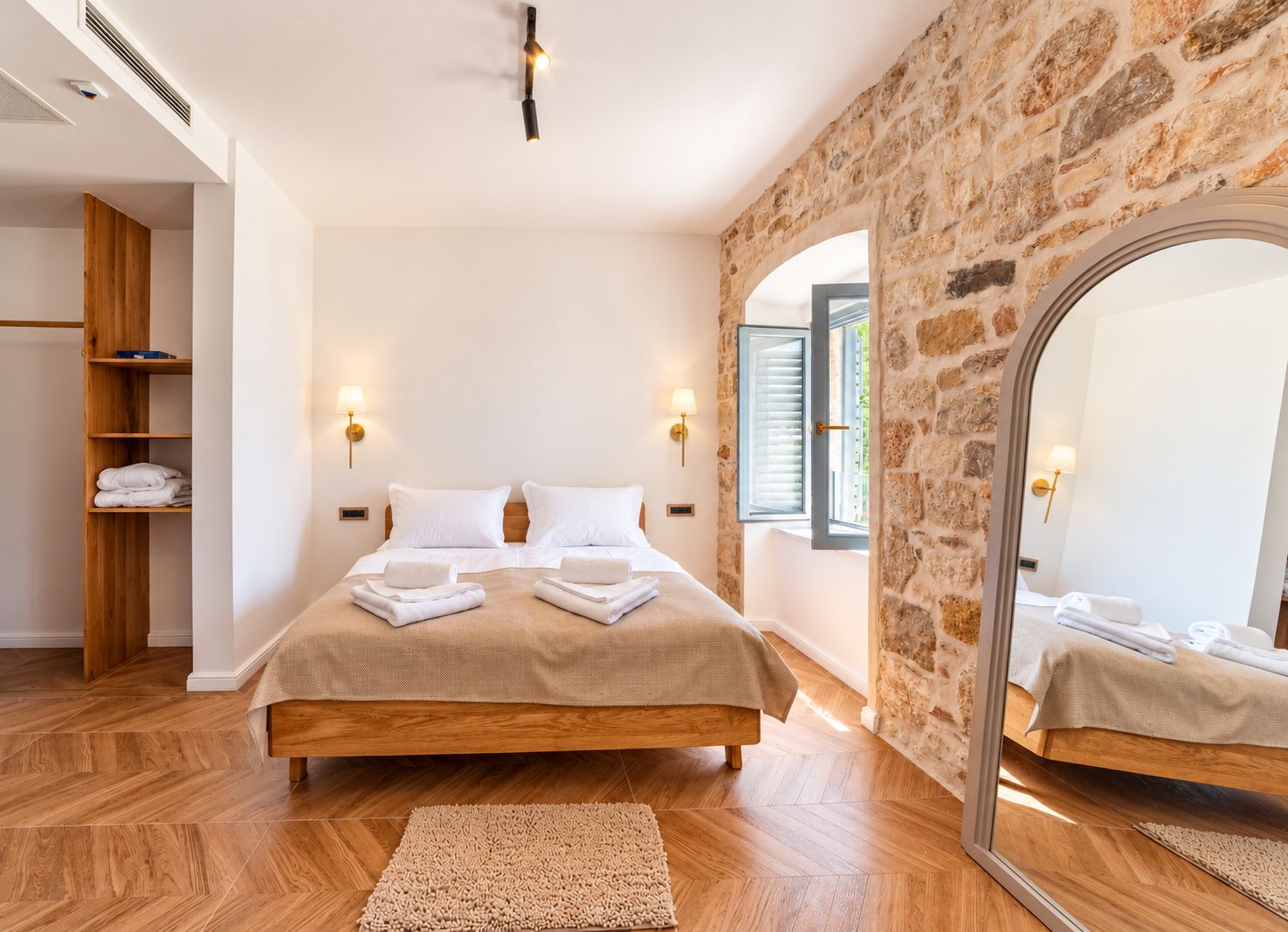 A bedroom with a large bed, wooden wardrobe, stone wall, and a mirror reflecting the room.