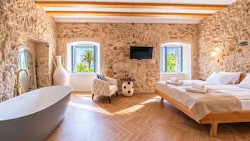 Une pièce avec un mur en pierre, une baignoire, un lit, un téléviseur et deux fenêtres donnant sur un paysage verdoyant.
