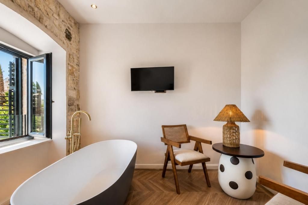 Un baño moderno con una bañera independiente, una silla de madera, una mesa redonda con una lámpara y un televisor de pantalla plana montado en la pared.