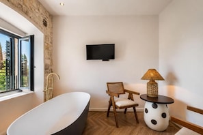 Un baño moderno con una bañera independiente, una silla de madera, una mesa redonda con una lámpara y un televisor de pantalla plana montado en la pared.