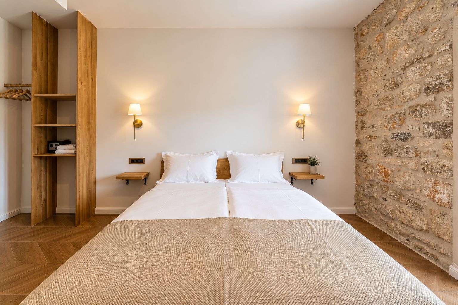 A double bed with a neutral-colored bedspread, two bedside tables with lamps, a wooden wardrobe, and a stone wall.