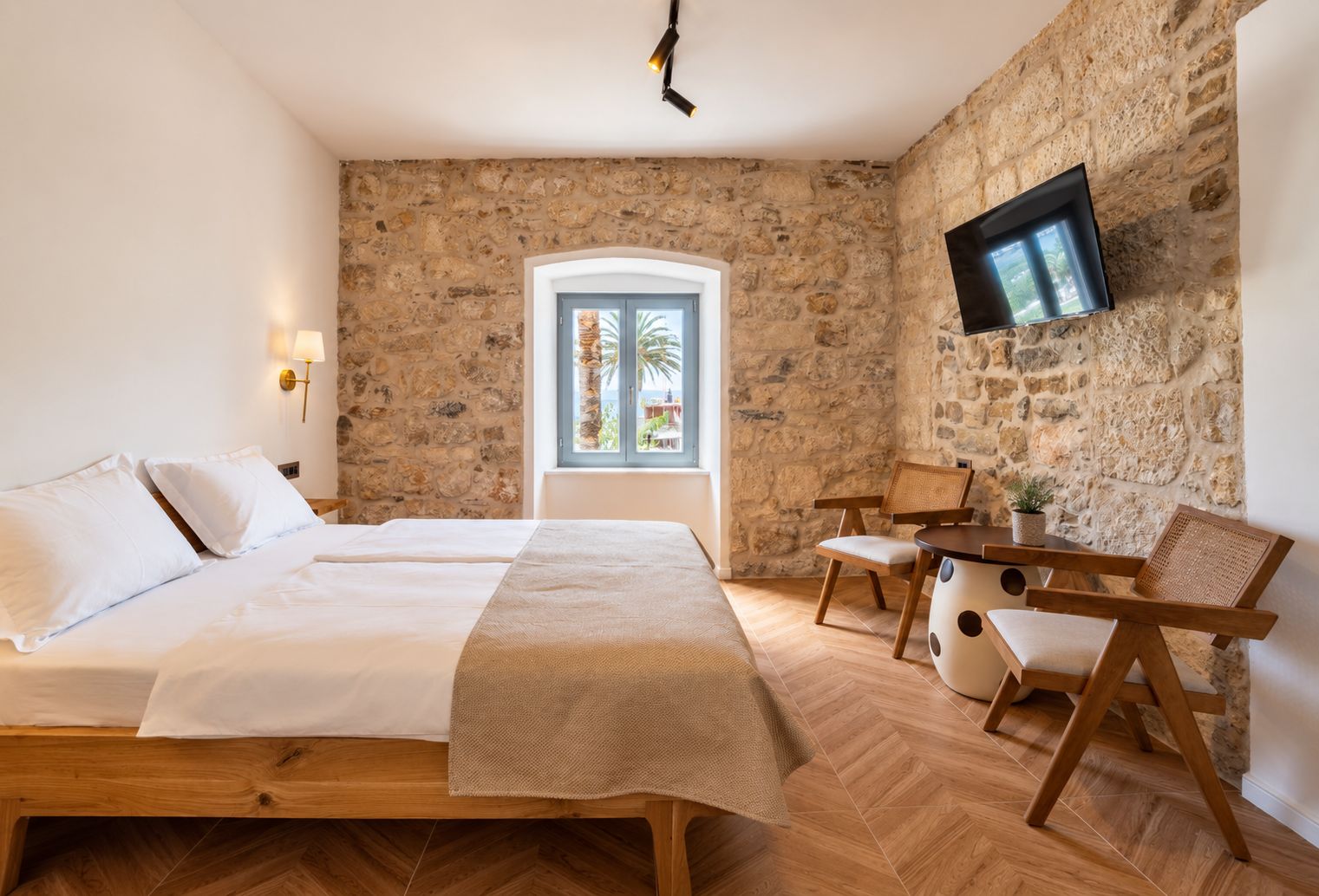 Une chambre avec un mur en pierre, un lit recouvert de draps blancs, un téléviseur fixé au mur et une fenêtre donnant sur des palmiers.