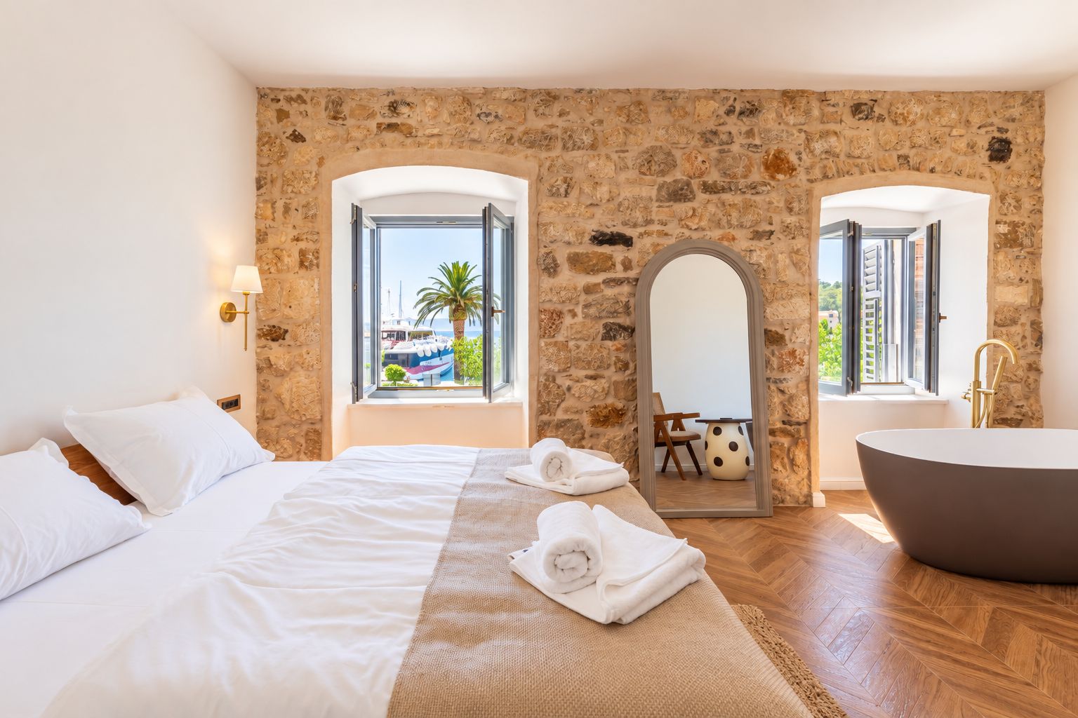 Une chambre d’hôtel avec un mur en pierre, un lit avec du linge de maison blanc, une grande fenêtre donnant sur les bateaux, une baignoire et un miroir.