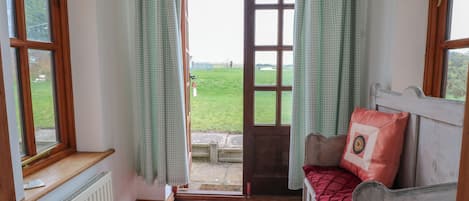 A room with a wooden bed, a red rug, a view of a green field through a door, and a window with a curtain.