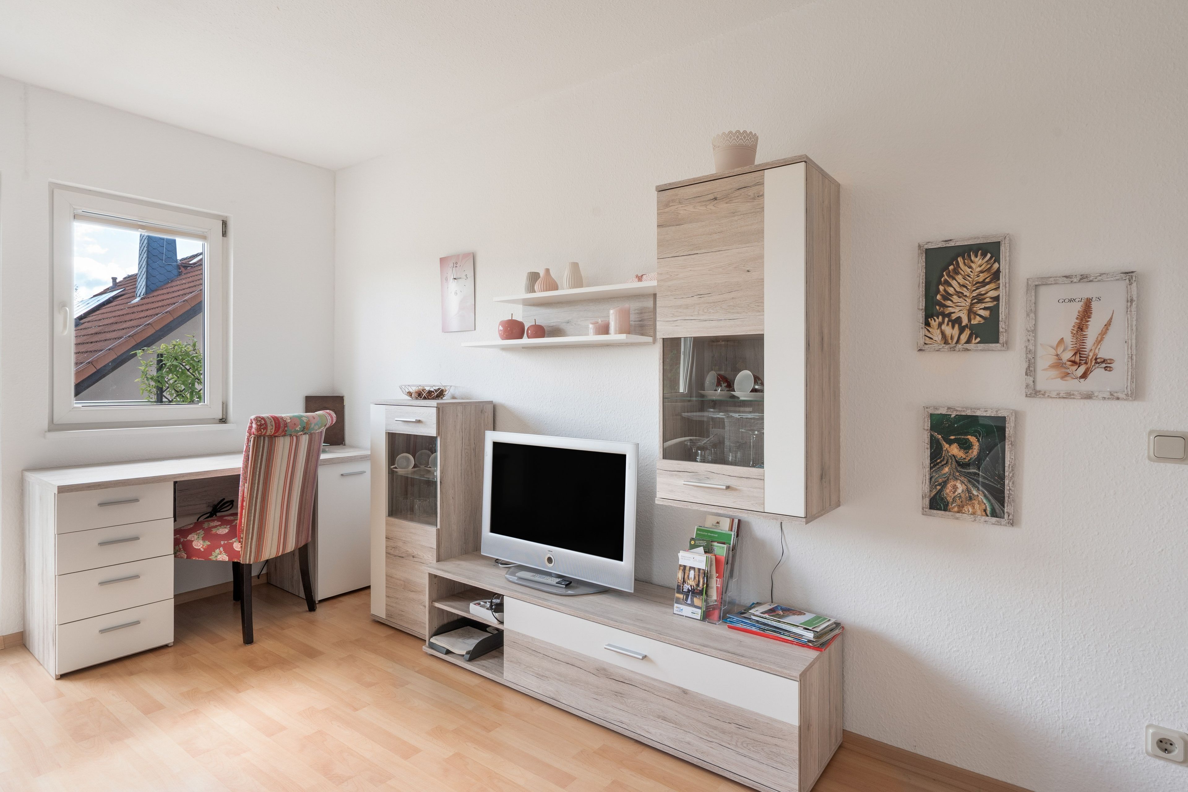 Ein modernes, helles Zimmer mit einem Holztisch, einem Fernseher und einem Fenster mit Blick auf ein Gebäude.