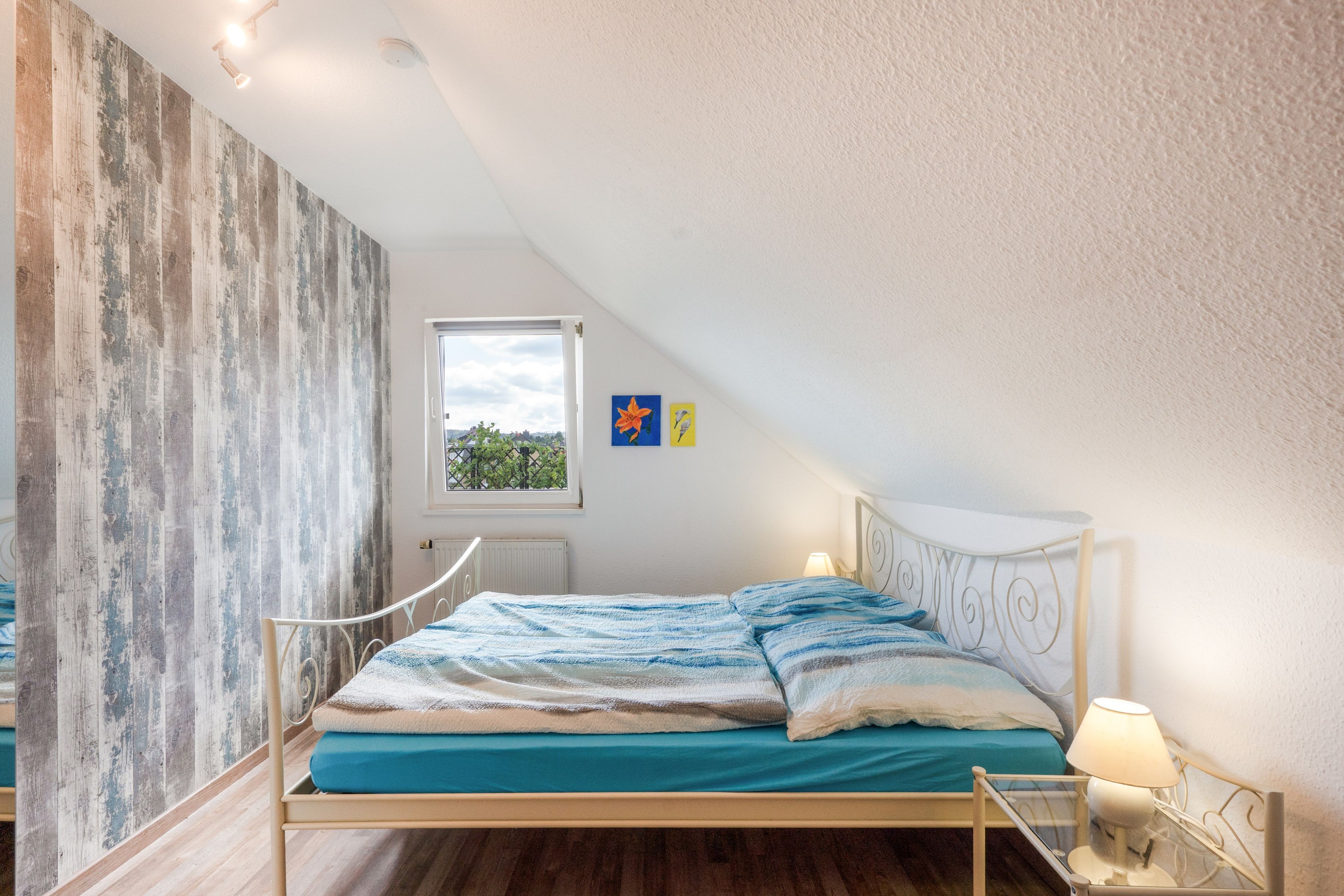 Une chambre avec un plafond en pente, un lit recouvert d’une couverture à rayures bleues et blanches, une fenêtre donnant sur la verdure et un mur à la texture bois.