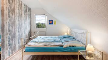 Ein Schlafzimmer mit geneigter Decke, einem Bett mit blau-weiß gestreifter Bettdecke, einem Fenster mit Blick auf Grünflächen und einer Wand mit Holzstruktur.