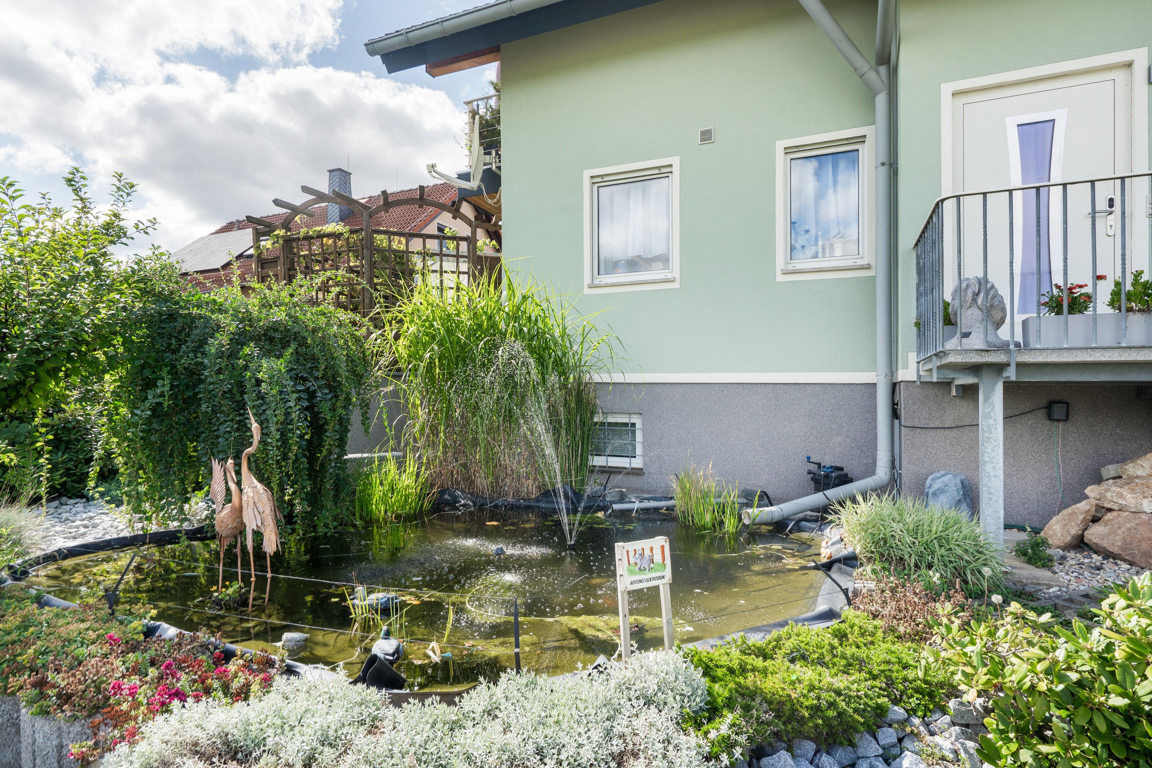 Un edificio residencial con un estanque en el jardín, una pérgola de madera y un balcón con plantas en macetas.