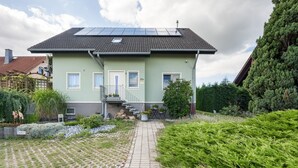 Une maison à deux étages, avec un toit recouvert de tuiles, des panneaux solaires et une allée pavée menant à l’entrée.