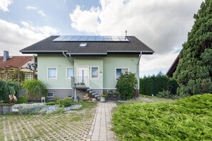 Une maison à deux étages, avec un toit recouvert de tuiles, des panneaux solaires et une allée pavée menant à l’entrée.