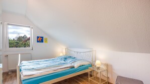 Un lit simple avec une tête de lit, une table de chevet avec une lampe et une fenêtre donnant sur un paysage verdoyant.