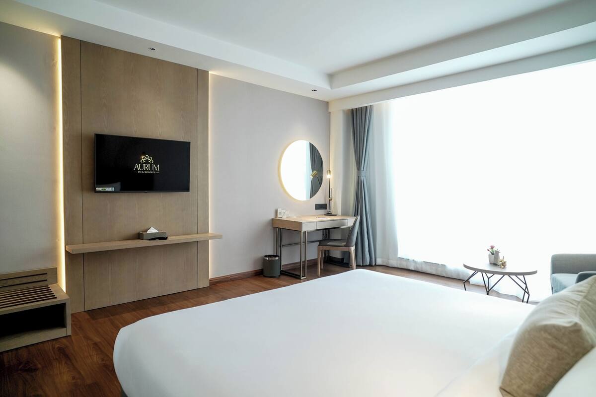 A modern hotel room with a large bed, a desk with a chair, a TV mounted on the wall, a round mirror, and a small table with a lamp.
