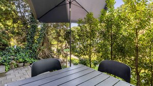 En terrasse med et bord og stole under en stor parasol, omgivet af grønne planter og træer.