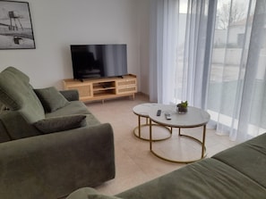 Ein Wohnzimmer mit einer Couch, einem Fernseher auf einem Holzgestell, zwei kleinen Tischen und einem Fenster mit durchsichtigen Vorhängen.