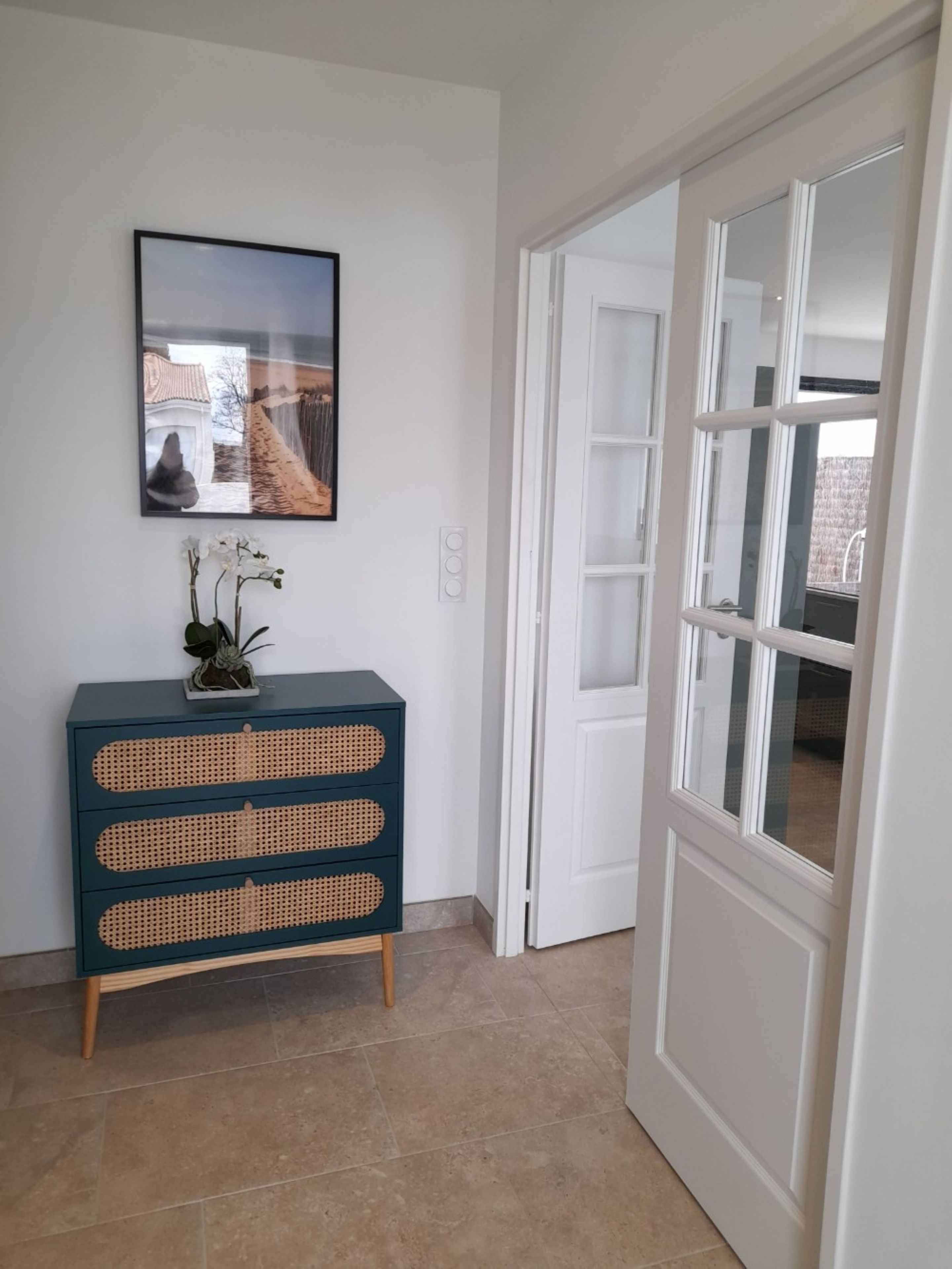 Un couloir avec une armoire bleu foncé à trois tiroirs, une plante en pot et un tableau encadré accroché au mur.