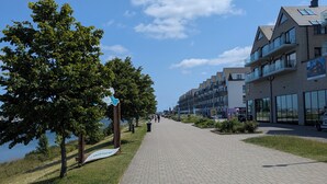 En asfalterad gångväg som löper längs med en vattenyta, med träd och moderna byggnader med balkonger på vardera sidan.
