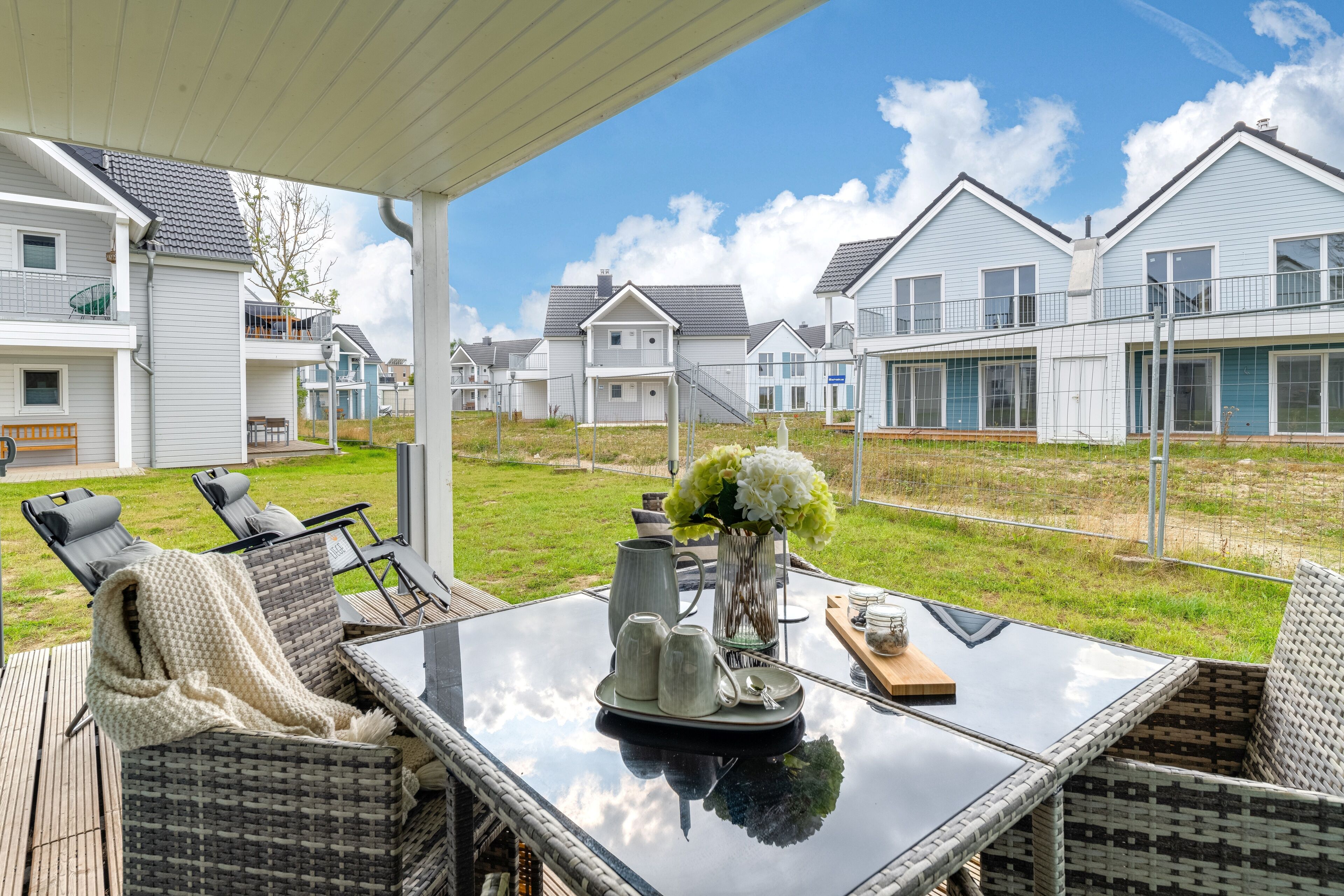 A covered patio area with wicker furniture, a glass table, and a view of modern houses.