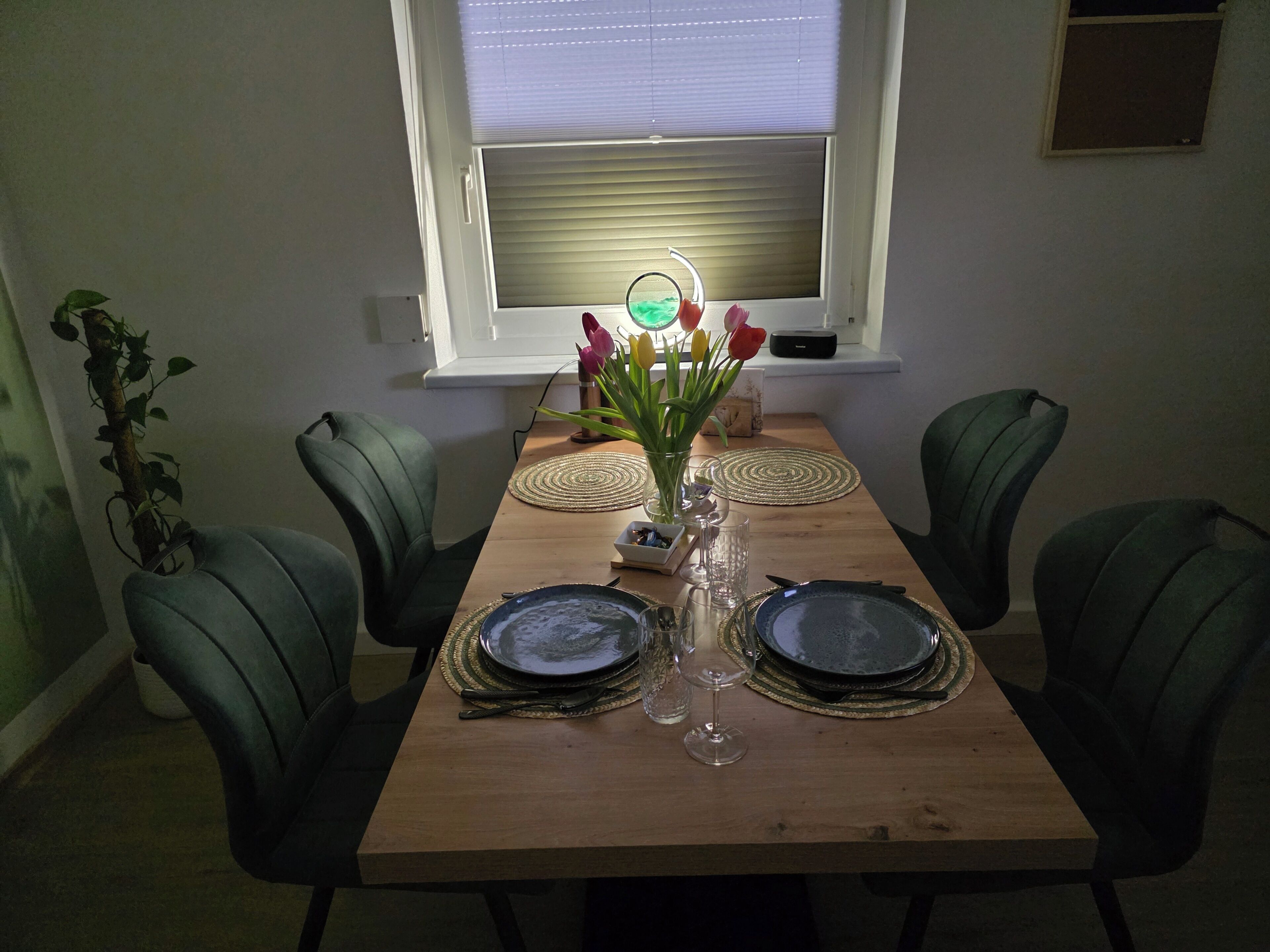 Une table à manger dressée pour six personnes, avec des chaises vertes, un vase rempli de fleurs et une fenêtre avec des stores.