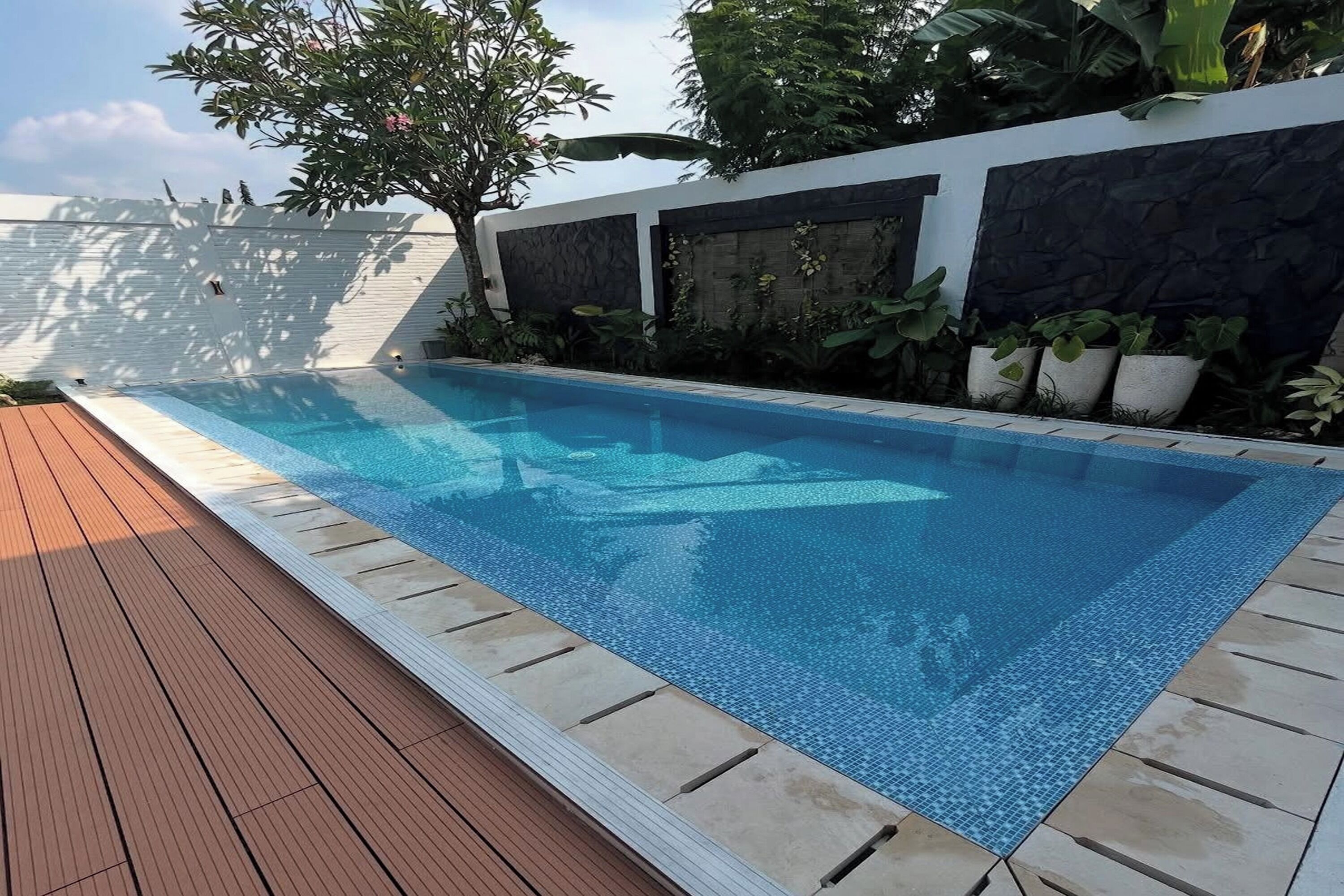 A rectangular swimming pool with a tiled edge, surrounded by a wooden deck and a white wall with a stone facade.