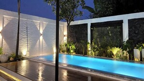 A swimming pool with a wooden deck, surrounded by plants and a white wall.