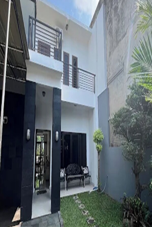 A two-story building with a balcony, a door, and a small garden area with a bench. - Mega Jaya Villa (Depok)