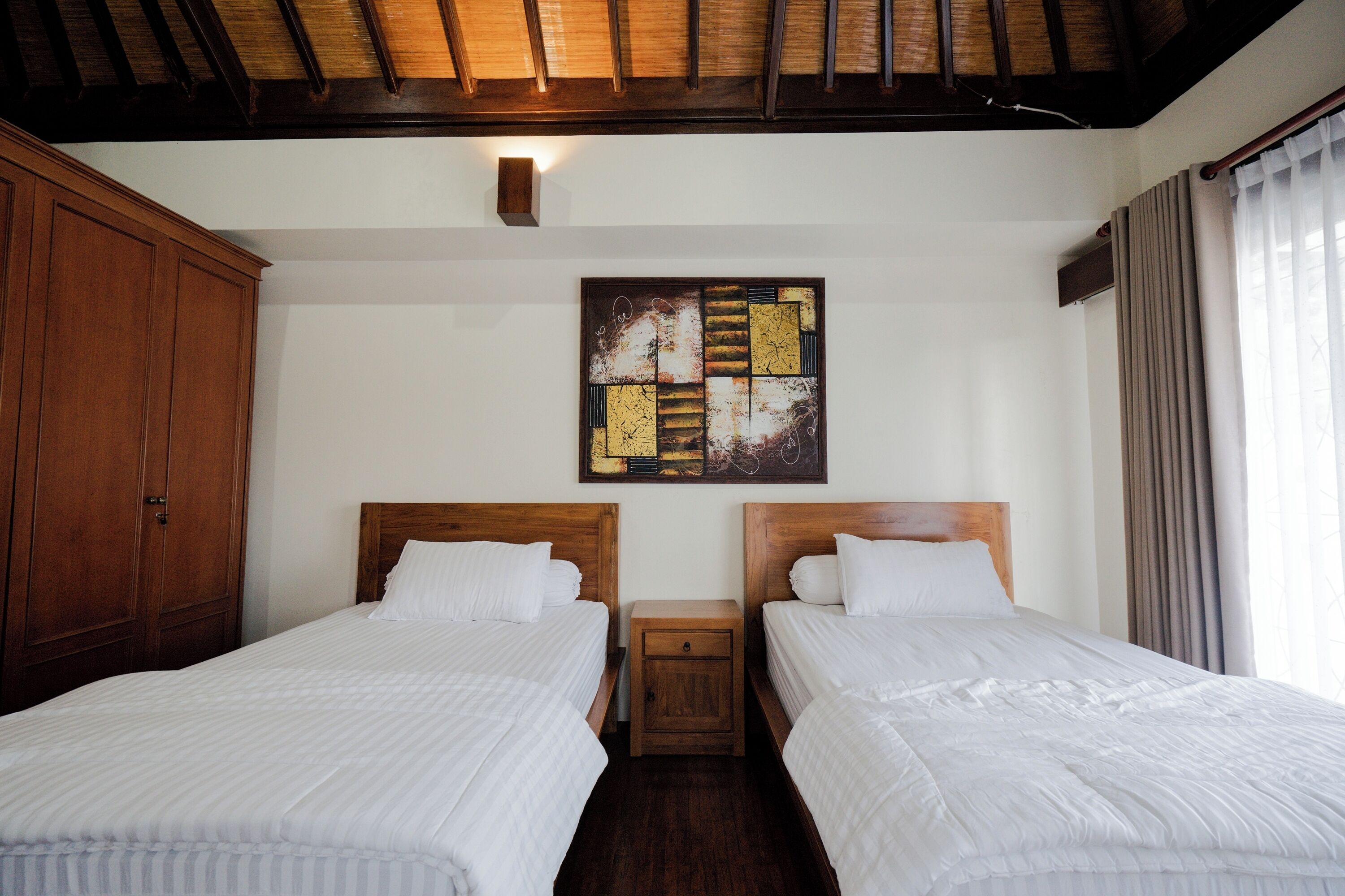 Habitación de hotel con dos camas, cabeceros de madera, una mesita de noche, una obra de arte colgada en la pared y una ventana con cortinas.